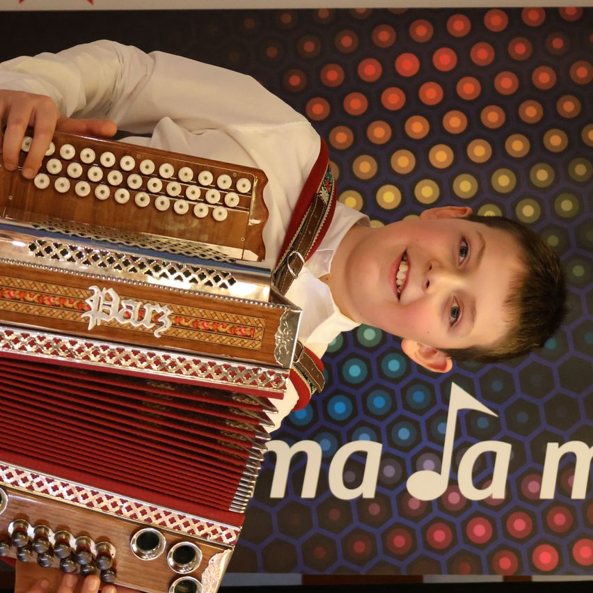 A young boy is playing an accordion, smiling, and holding it close to his face. The background is a colorful pattern.