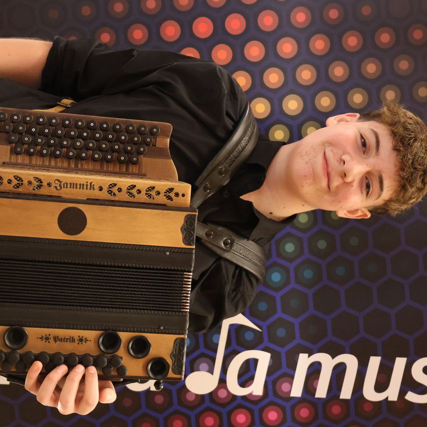 A young man is smiling and holding an accordion, wearing a black shirt. He is standing in front of a colorful hexagonal pattern background with the word 'La Mus'.