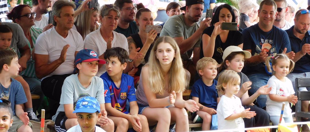 Eine Gruppe von Menschen beobachtet eine Veranstaltung im Freien, auf Gras und Tribünen sitzend. Kinder und Erwachsene klatschen. Bunte Wimpel schmücken die erste Reihe.