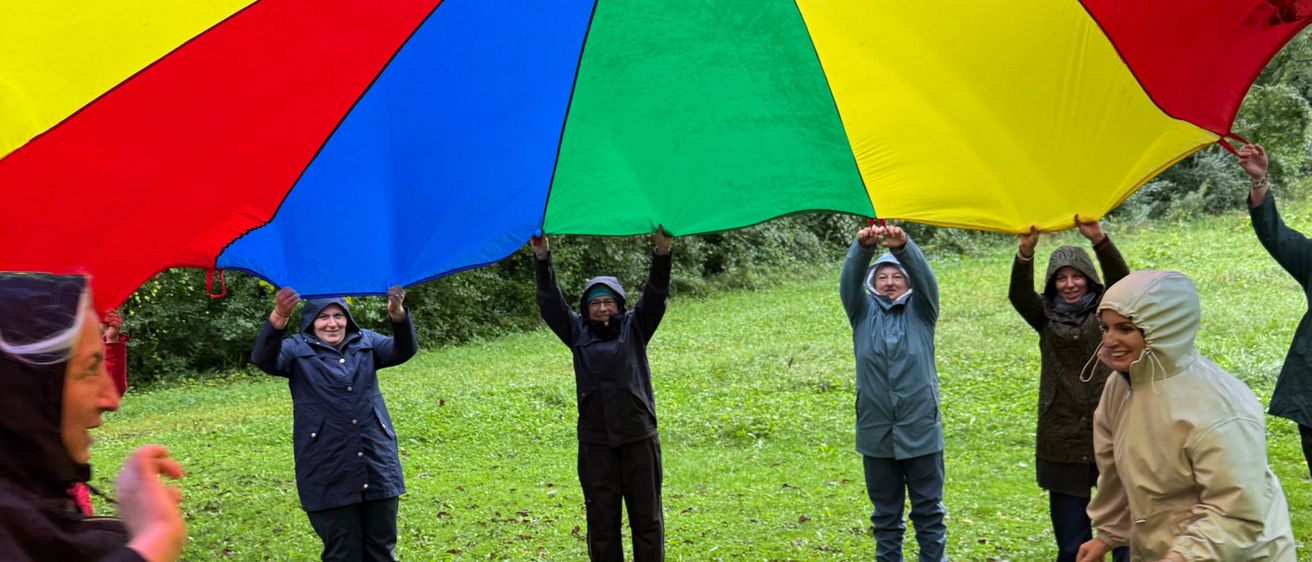 Eine Gruppe von Menschen hält einen großen, bunten Regenschirm, während sie auf einem Grasfeld stehen. Sie sind in Regenmänteln gekleidet und scheinen Spaß zu haben.