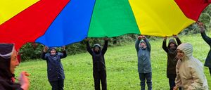 Eine Gruppe von Menschen hält einen großen, bunten Regenschirm, während sie auf einem Grasfeld stehen. Sie sind in Regenmänteln gekleidet und scheinen Spaß zu haben.