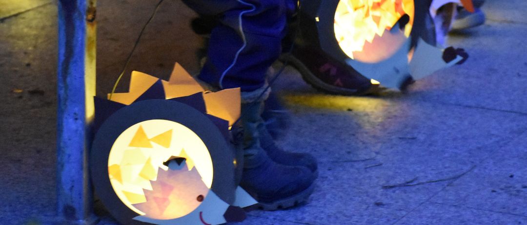 Ein Kind hält eine Laterne in Form eines Igels mit beleuchtetem Inneren und sitzt neben einer anderen Person mit ähnlicher Laterne in einem schwach beleuchteten Bereich.