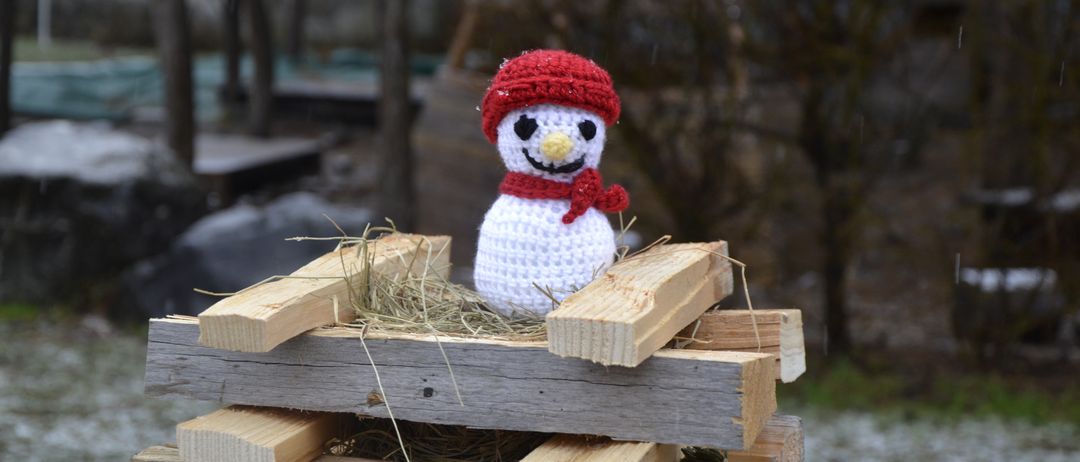 Ein gehäkelter Schneemann ruht auf einem Stapel Holzbalken, trägt einen roten Hut und Schal. Er sitzt auf Heu mit kahlen Bäumen und Schnee im Hintergrund.