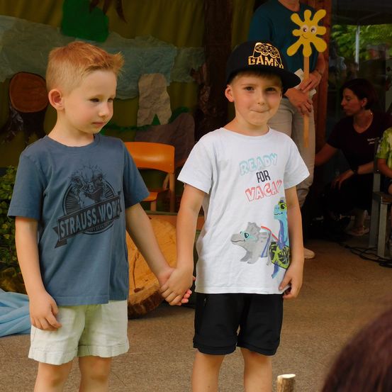 Zwei junge Jungen halten Händchen, einer trägt ein graues T-Shirt und der andere ein weißes. Sie stehen in einem Raum mit Holzboden.