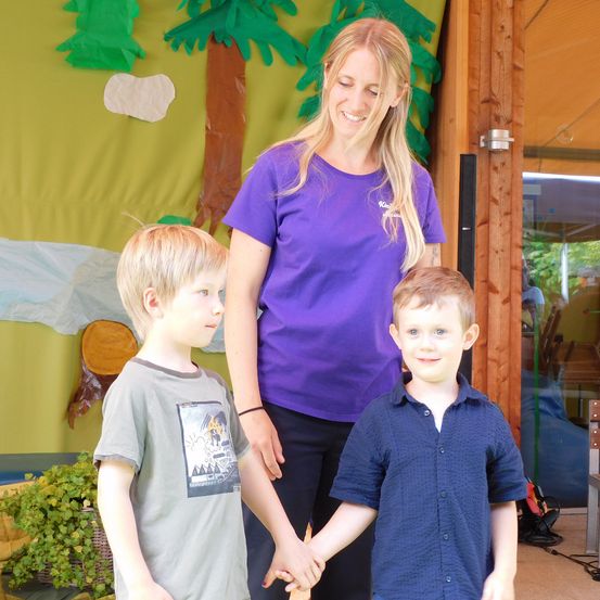 Eine Frau mit langen blonden Haaren lächelt, während sie die Hände von zwei Jungen hält, vor einer Wandmalerei.