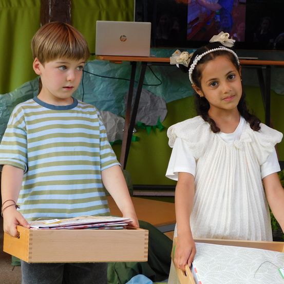 Ein Junge und ein Mädchen stehen zusammen und halten Holzschubladen, mit einem Laptop auf einem Tisch und einem Fernseher dahinter. Der Junge trägt ein gestreiftes Shirt, und das Mädchen hat eine Stirnband und ein weißes Kleid.