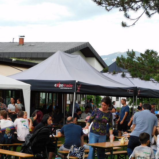 Eine Gruppe von Menschen isst bei einer Outdoor-Veranstaltung unter einem großen Zelt. Das Zelt ist schwarz mit dem Logo expotrade. Eine Frau steht in der Nähe eines Tisches mit Essen und Getränken. Im Hintergrund sitzen Menschen an Tischen und Gebäude mit Blick auf den Berg.