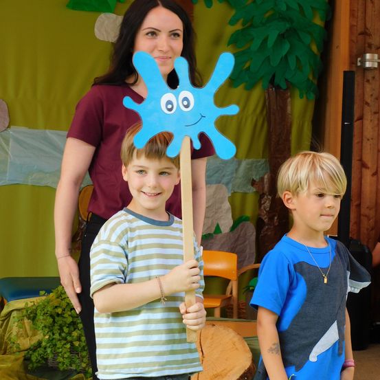 Eine Frau steht hinter zwei lächelnden Jungen, einer hält eine blaue Tropfenpuppe, vor einem Hintergrund mit einem Baum und Pflanzen.