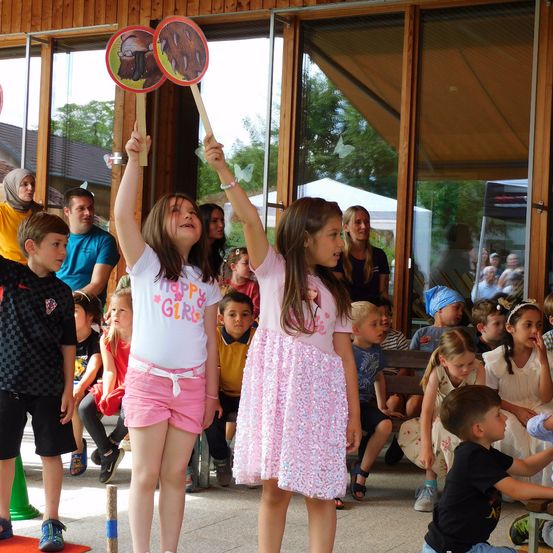 Zwei Mädchen in rosa Kleidern halten ein Schild mit einem Bären hoch, während eine Menschenmenge von Kindern und Erwachsenen zusieht. Ein Junge reicht nach einer Mädchen vor ihm.