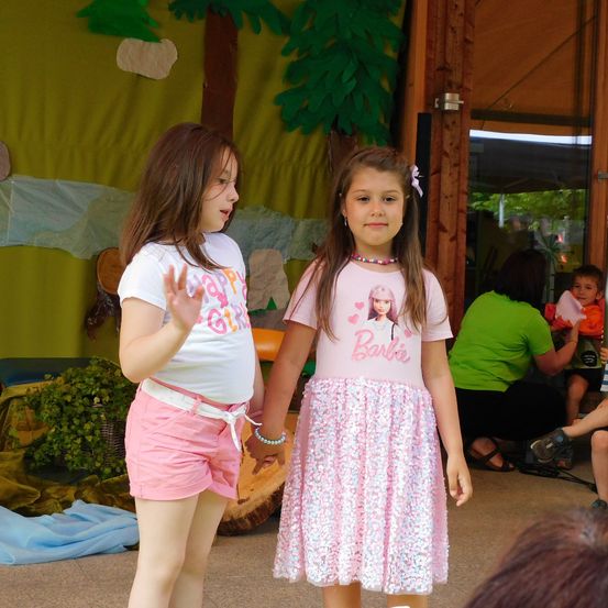 Zwei junge Mädchen stehen in einem Spielbereich mit Dekorationen hinter ihnen. Das Mädchen auf der linken Seite trägt ein weißes Shirt mit pinken Shorts. Das Mädchen auf der rechten Seite trägt ein rosa Barbie-thematisches Kleid. Im Hintergrund sitzt eine Person und ein Kind sitzt auf einem Schoß.