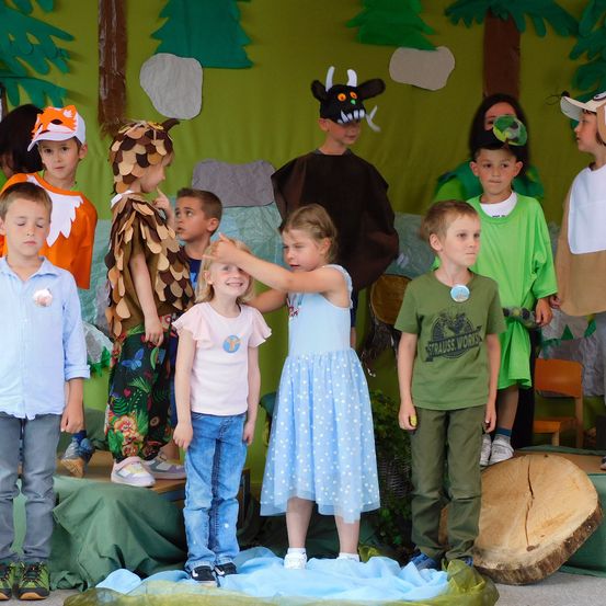 Mehrere Kinder stehen in Kostümen auf der Bühne für ein Theaterstück. Ein Kind in einem Fuchskostüm und ein anderes in einem Wolfskostüm sind darunter. Sie befinden sich in einer grünen, waldähnlichen Umgebung.