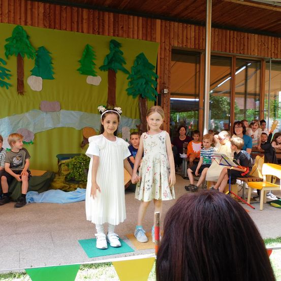 Zwei junge Mädchen in weißen Kleidern stehen auf einer Bühne vor einem grünen Hintergrund mit Bäumen. Hinter ihnen sitzen mehrere Kinder in Stühlen und andere stehen und beobachten die Aufführung.