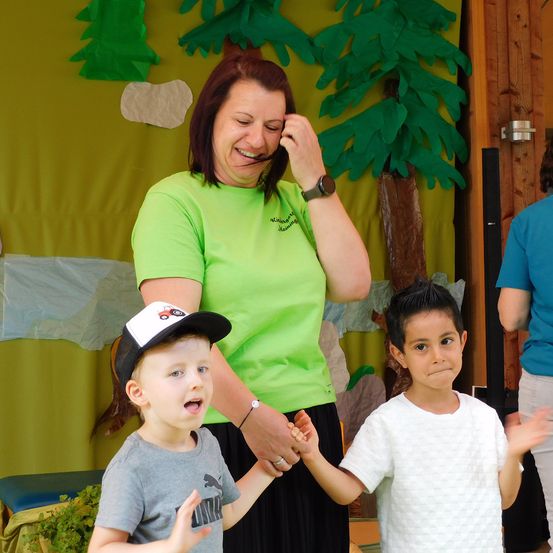 Eine Frau in einem grünen T-Shirt hält die Hände von zwei Jungen, einer in einer Mütze und der andere in einem weißen T-Shirt, alle stehen vor einem grünen Hintergrund.