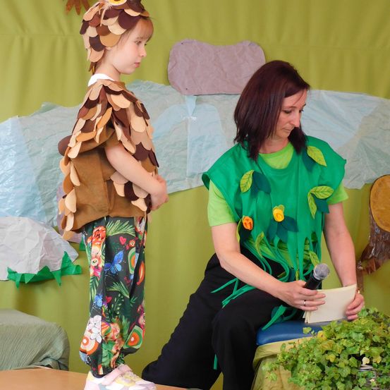 Ein Kind in einem Waldtierkostüm und eine erwachsene Frau in einem Blattkostüm sitzen auf einer Bühne und halten ein Mikrofon. Sie stehen vor einem grünen Hintergrund.