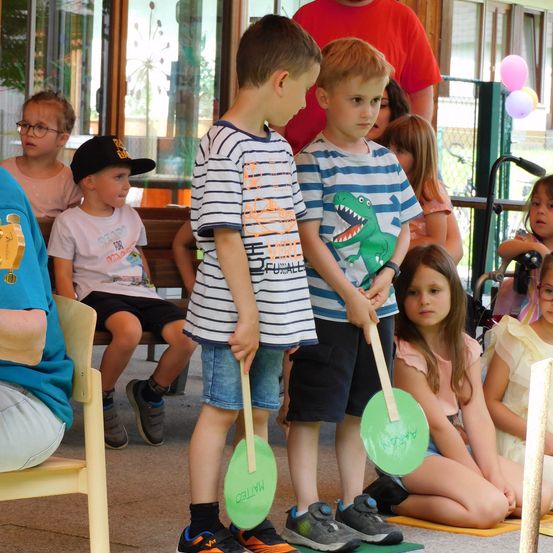 Eine Gruppe von Kindern steht in einer Reihe mit grünen Namensschildern, wahrscheinlich an einer Schulaktivität teilnehmend. Sie stehen vor einer Bank mit einer sitzenden Person. Im Hintergrund sind Luftballons und ein Zaun zu sehen.