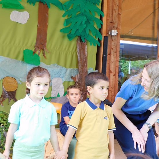 Drei Kinder halten Händchen, eine lächelnde Frau in der Nähe. Sie stehen vor einer bemalten Kulisse mit Bäumen und einer Landschaft.