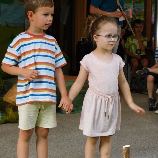 Ein junger Junge und ein Mädchen halten sich an den Händen in einem Raum, in dem andere auf dem Boden sitzen. Das Mädchen trägt eine Brille und ein rosa Kleid, während der Junge ein gestreiftes T-Shirt trägt. Hinter ihnen sitzen Leute auf dem Boden, und einer hält einen Stock.