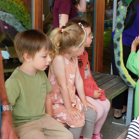 Ein Junge und zwei Mädchen sitzen auf einer Bank und schauen sich etwas vor sich an. Das Mädchen in der Mitte trägt eine Perlenkopfband. Dahinter steht eine Person mit einem Tattoo am Arm.