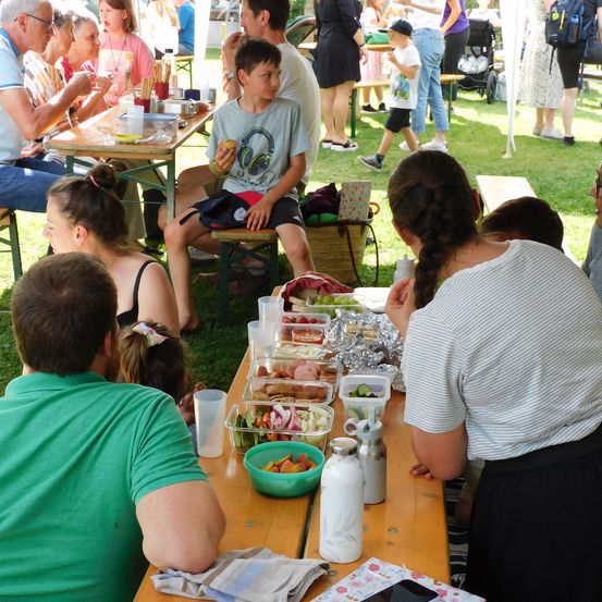 Mehrere Familien essen im Freien an einem Picknicktisch. Eine Frau in einem weiß gestreiften Oberteil steht neben dem Tisch, und ein Junge in einem grauen T-Shirt isst mit den Händen.