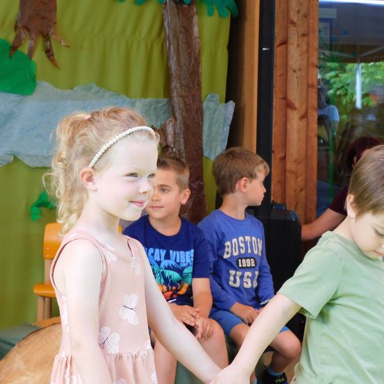 Ein kleines Mädchen mit einem rosa Kleid und einer Stirnband lächelt, während es die Hand eines Jungen hält. Hinter ihnen sitzen drei Jungen auf einer Bank. Die Umgebung hat einen grünen Hintergrund mit Baumausschnitten und einer Holzwand.