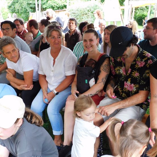 Eine Familie mit kleinen Kindern und Erwachsenen in einem Garten, einige mit Hüten, sitzend und stehend, mit Bäumen im Hintergrund.