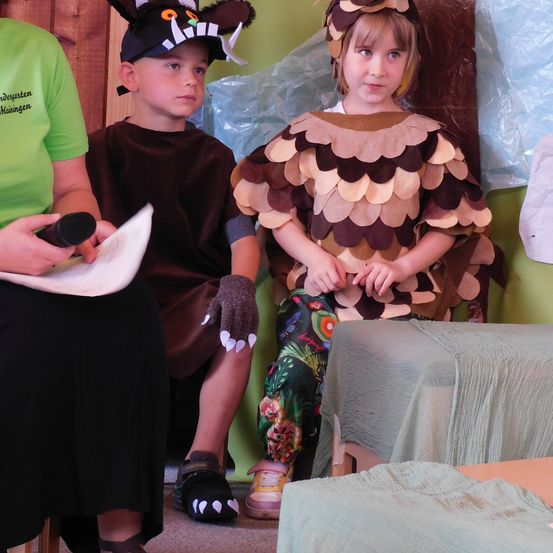 Zwei Kinder in Kostümen, eines als Eule und das andere als Fuchs, sitzen in einem Raum mit einer grünen Wand. Eine Person mit einem Mikrofon steht vor ihnen.