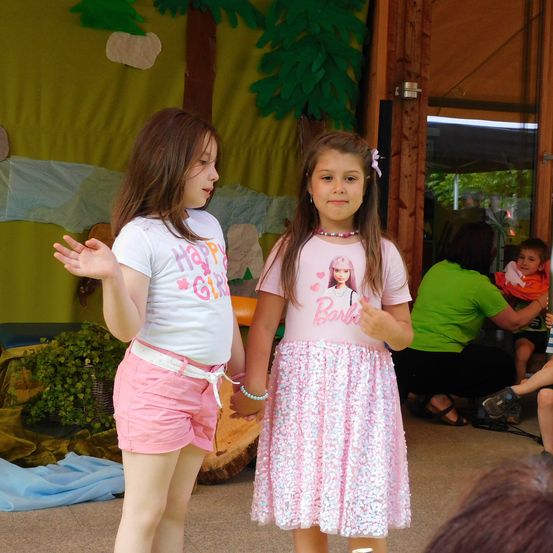 Zwei junge Mädchen stehen nebeneinander, eines trägt ein 'Happy Girl' T-Shirt und das andere ein 'Barbie' T-Shirt. Beide sind in rosa Röcken gekleidet und schauen auf etwas vor ihnen. Dahinter sitzt eine Frau in einem grünen Shirt mit einem Baby.