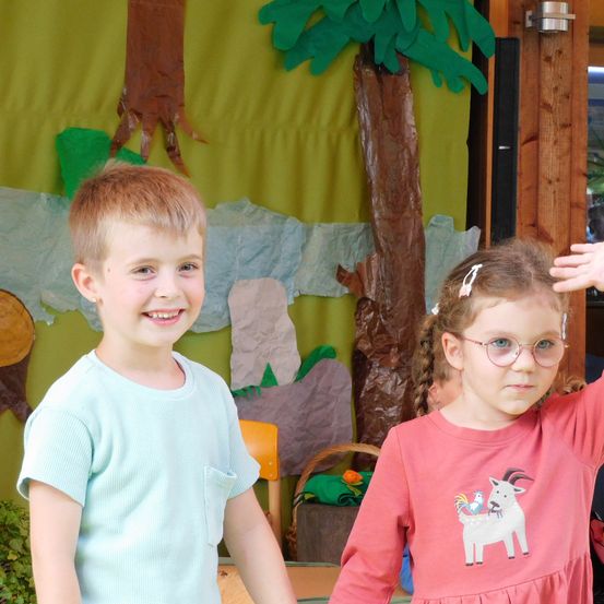 Zwei Kinder stehen in einem Klassenzimmer mit einem Dschungel-Hintergrund. Ein Junge mit einem mintgrünen Shirt winkt, während ein Mädchen mit Brille lächelt. Dahinter sind eine Holzstruktur und ein Korb zu sehen.