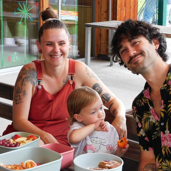Eine dreiköpfige Familie sitzt lächelnd an einem Tisch im Freien. Die Frau hat Tattoos an den Armen und der Mann trägt ein Blumenhemd. Ein Kleinkind sitzt zwischen ihnen und hält ein Stück Obst. Der Tisch ist mit einer Schüssel Obst und einem Behälter mit Gebäck gedeckt.