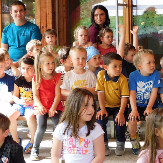 Eine Gruppe von Kindern und zwei Erwachsenen sitzen auf einer Bank, wobei einige Kinder die Hände heben. Die Kinder tragen Freizeitkleidung, und einige tragen eine Brille. Die Erwachsenen stehen hinter den Kindern, und einer von ihnen hält eine Kamera. Der Hintergrund ist unscharf, mit einer Holzwand und einer Glastür.