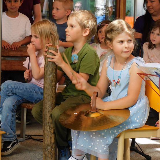 Mehrere Kinder sitzen in einer Reihe mit Musikinstrumenten, darunter eine Becken. Ein Junge hält einen Stock und das Becken, während ein Mädchen neben ihm ein Notenbuch hält. Dahinter sitzen weitere Kinder und beobachten.