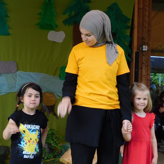 Eine Frau in einem gelben T-Shirt und einem Kopftuch steht zwischen zwei Mädchen, eines hält ihre Hand. Das Mädchen auf der linken Seite trägt ein Pikachu-T-Shirt, während das Mädchen auf der rechten Seite ein rotes Kleid trägt. Dahinter befindet sich ein grüner Hintergrund mit Papierbäumen.