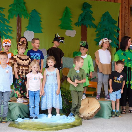 Kinder in verschiedenen Kostümen stehen auf einer Bühne mit einer waldthematischen Kulisse. Ein Baumstamm liegt vor ihnen auf dem Boden.
