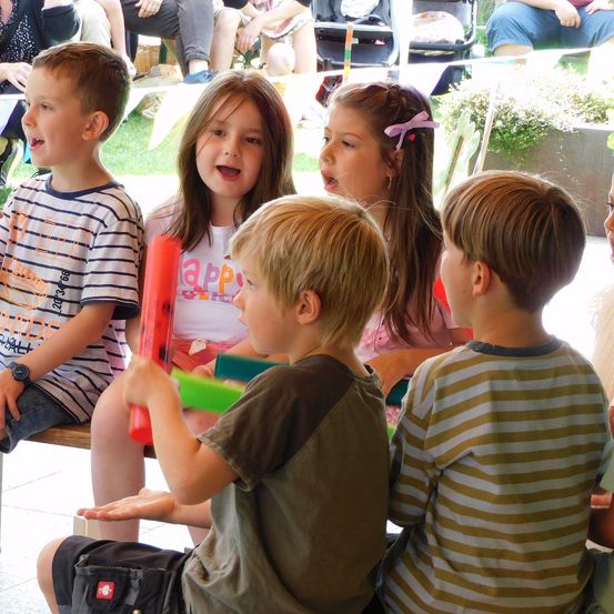 Fünf Kinder sitzen auf einer Bank und singen und spielen Instrumente. Zwei Mädchen sind in der Mitte, und drei Jungen umgeben sie. Sie befinden sich in einem grünrasigen Außenbereich.