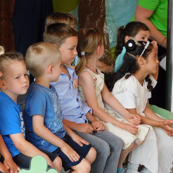 Eine Gruppe von Kindern sitzt zusammen auf einer Bank. Ein Mädchen mit langen Haaren trägt ein weißes Kleid, während ein Junge neben ihr ein blaues Shirt trägt. Im Hintergrund beobachtet eine Frau in einem grünen Shirt sie.