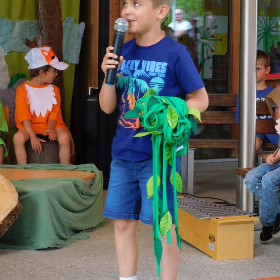 Ein Junge hält ein Mikrofon und singt, mit grünen Blättern um die Taille. Hinter ihm sitzt ein anderer Junge in einem Fuchskostüm auf einem Baumstumpf. Ein Xylophon liegt auf dem Boden.