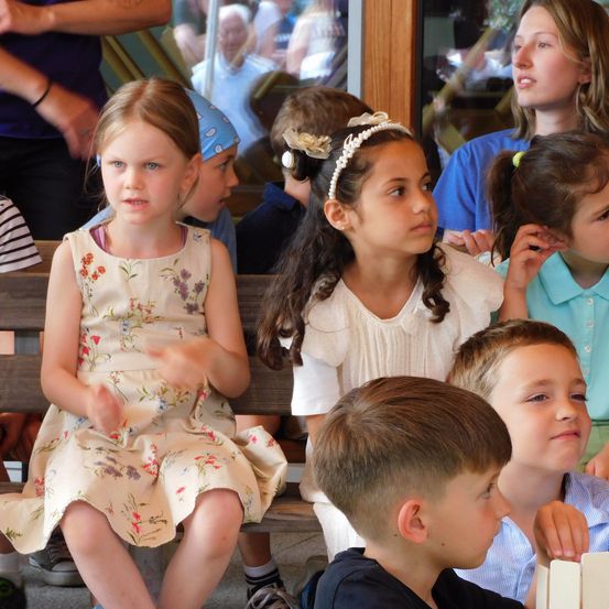 Eine Gruppe von Kindern sitzt auf einer Bank, wobei ein Mädchen in einem Blumenkleid vorne sitzt. Dahinter sitzen andere Kinder, eines mit einem Stirnband. Der Hintergrund ist unscharf und zeigt weitere Kinder und Erwachsene.
