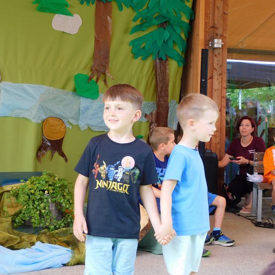 Zwei Jungen stehen vor einer Baumkulisse, einer trägt ein Ninjago-T-Shirt. Eine Frau sitzt in der Nähe, und ein weiteres Kind sitzt auf dem Boden.