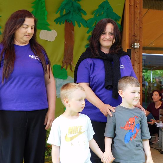 Zwei Frauen und zwei junge Jungen stehen vor einer Kulisse mit grünen Bäumen. Die Frauen tragen lila T-Shirts und die Jungen weiße und graue T-Shirts. Der Junge auf der rechten Seite hält die Hand des Jungen auf der linken Seite.