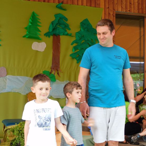 Ein Mann in einem blauen T-Shirt steht mit zwei Jungen vor einer Kulisse mit Ausschnitten von Bäumen und einem Berg.