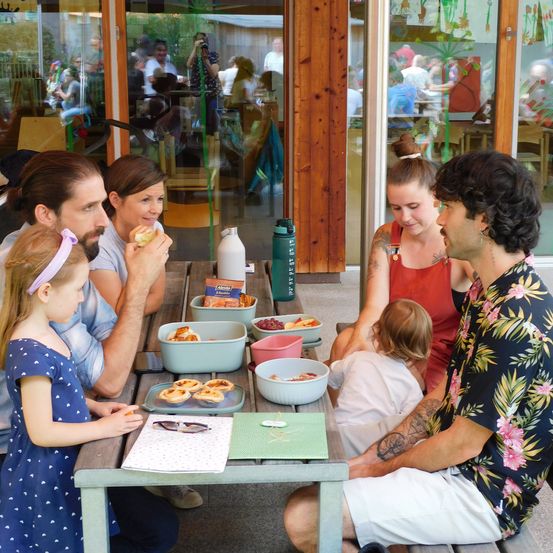 Eine Familie sitzt an einem Tisch in einem Restaurant und isst. Auf dem Tisch stehen ein Teller, eine Schüssel und eine Schachtel. Die Familie besteht aus einem Mann, zwei Frauen und einem Kind.
