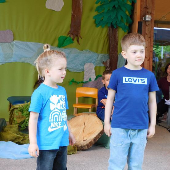 Zwei Kinder, ein Mädchen mit einem Pferdeschwanz und ein Junge in einem blauen Levis-T-Shirt, stehen in einem Raum mit einem grünen Hintergrund. Ein Baumstamm und ein orangefarbener Stuhl sind hinter ihnen.