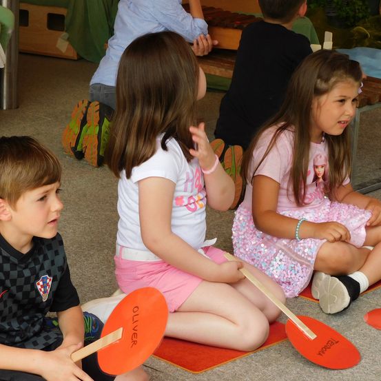 Kinder sitzen auf dem Boden in einem Klassenzimmer. Zwei Mädchen halten Holzstäbe. Ein Junge schaut etwas an, während er ein Schild mit dem Namen Oliver hält.