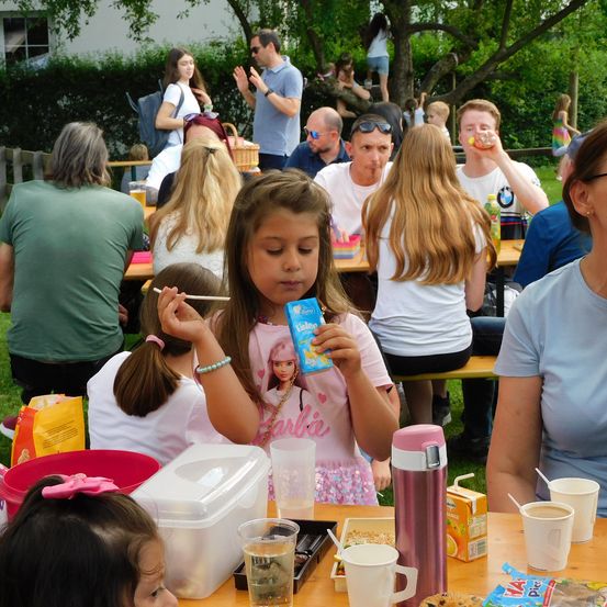 Eine Gruppe von Menschen sitzt an Tischen im Freien, manche essen. Ein junges Mädchen in einem rosa Barbie-Shirt hält eine Müslischachtel. Becher, ein Thermos und Essen stehen auf den Tischen. Bäume und ein Gebäude sind im Hintergrund.