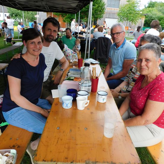 Eine vierköpfige Familie sitzt an einem langen Holztisch im Freien. Der Tisch ist mit Essen und Getränken beladen. Ein Baby ist in einem Kinderwagen in der Nähe.