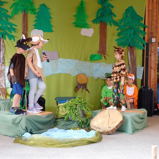 Vier Kinder in Kostümen stehen auf einer Bühne mit einem Waldhintergrund. Eines hält ein Mikrofon, während andere auf Baumstämmen sitzen oder stehen. Die Bühne hat grünen Stoff, der den Boden und die Bäume bedeckt.