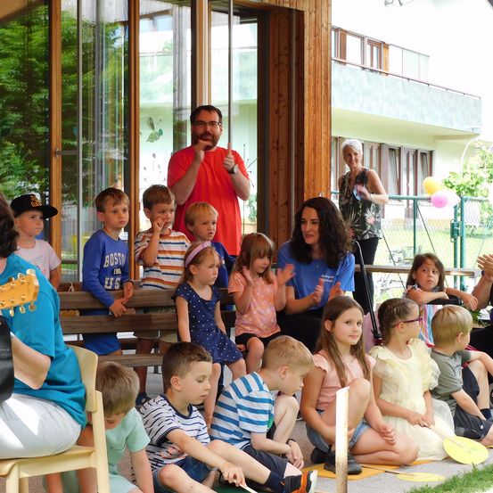 Eine Gruppe von Kindern sitzt auf einer Bank und beobachtet einen Mann, der Gitarre spielt. Dahinter steht eine Frau mit erhobenen Händen. Im Hintergrund befindet sich ein Gebäude mit Balkon und einem Zaun mit Luftballons.