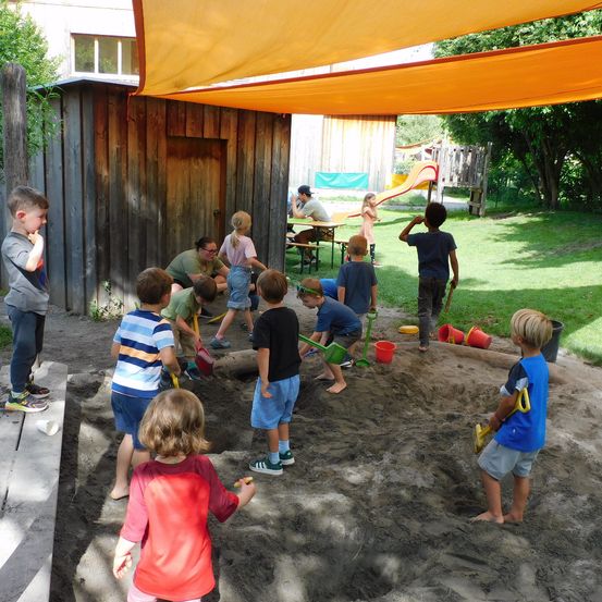 Mehrere Kinder spielen mit Spielzeug in einem Sandkasten unter einem Baldachin, ein Erwachsener beobachtet in der Nähe.