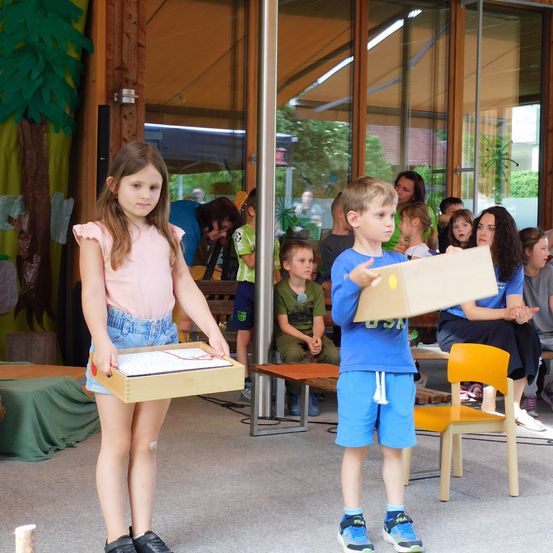 Zwei Kinder stehen vor einem Publikum mit Kisten in der Hand, eines schaut das andere an, umgeben von anderen Kindern und Erwachsenen.
