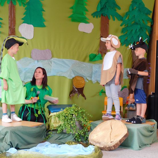 Drei Kinder in Kostümen stehen auf der Bühne mit einer Kulisse von Bäumen und einem Fluss. Eine Frau in einem grünen Kostüm hält ein Mikrofon und scheint sie zu dirigieren.